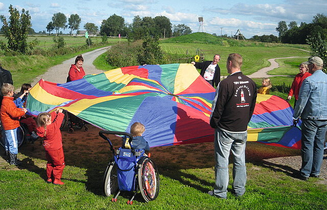 Eine Gruppe von Erwachsenen und Kindern mit und ohne Rollstühlen spiegeln mit einem bunten Tuch auf einer Wiese