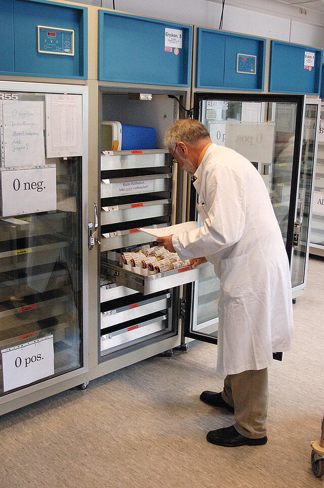 Fotografie eines Mannes mit weißem Kittel vor einem Kühlschrank mit Blutkonserven.