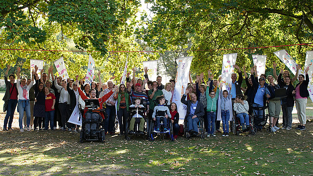 Gruppenfoto von winkenden Menschen unterschiedlichen Alters, mit und ohne Behinderung.