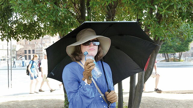 Eine Frau mit Handschuhen und Sonnenbrille steht im Freien unter einem großen Schirm.